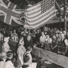 Crowd watches man hammer boat plank underneath the British and American flags