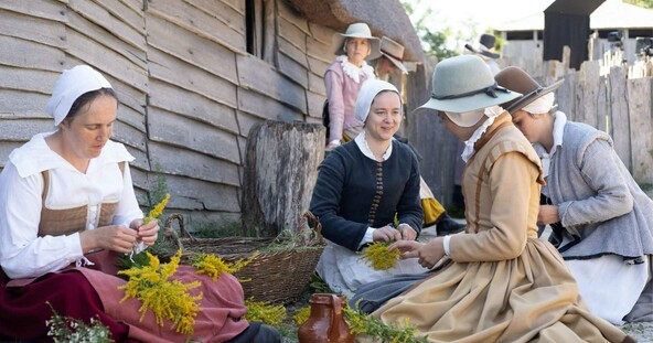 A group of seated Pilgrim women gather flowers outside of a grey house