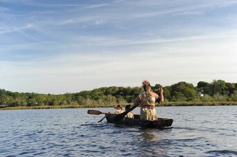 Man wearing regalia paddles his two daughters over water in a mishoon