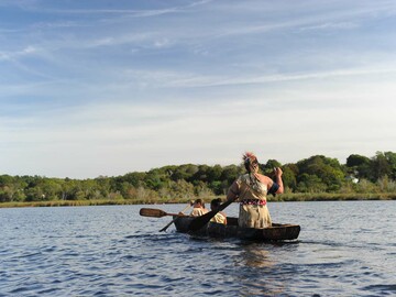 Man wearing regalia paddles his two daughters over water in a mishoon