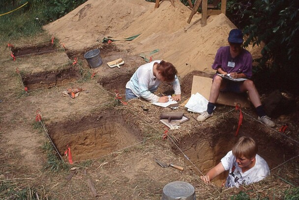 Three archaeologists methodically record their work. Multiple large, square shaped holes in the ground are present.