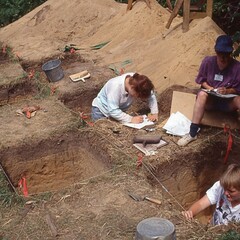 Three archaeologists methodically record their work. Multiple large, square shaped holes in the ground are present.