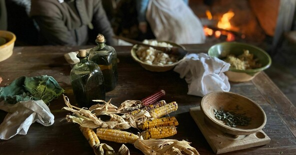 An array of food items and serve wear on a wooden table. Two pilgrims sit behind the table.