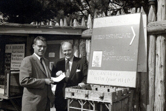 Two men in business suits stand in front of a sign and model of Plymouth Colony’s Fort Meeting House
