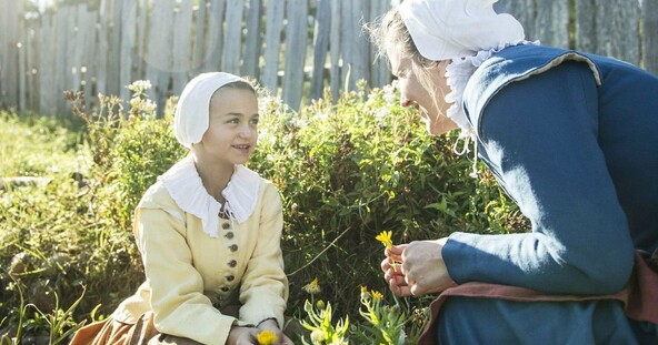 A pilgrim mother and daughter crouch in a garden while talking and picking flowers