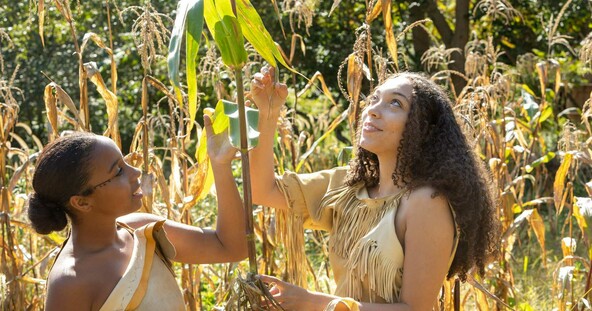 Two young women dressed in regalia look at growing corn