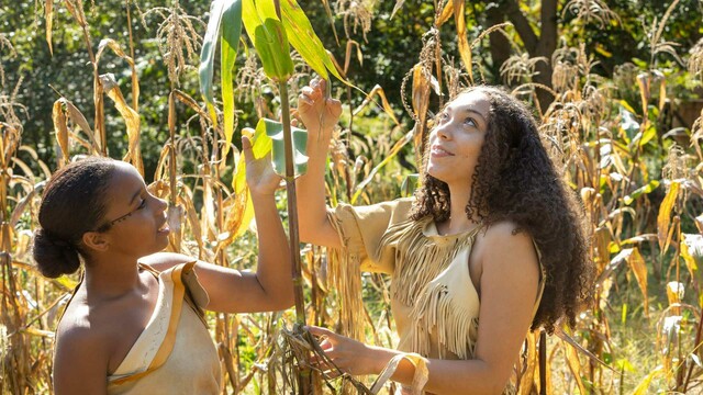 Two young women dressed in regalia look at growing corn