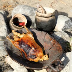 Roast chicken in a wooden bowl is set over stones. Cooking pots are in the background.