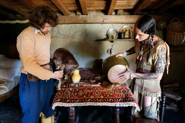 A pilgrim man and Wampanoag man inspect objects for trade in a home interior