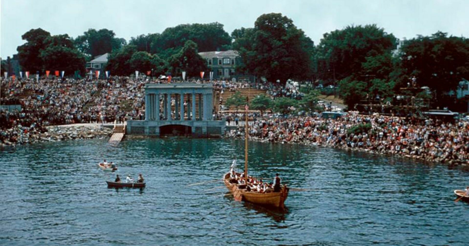 A shallop is rowed towards Plymouth Rock while a crowd of spectators looks on