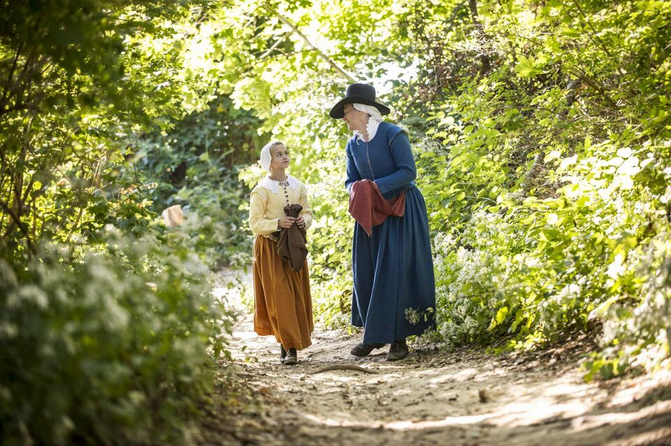 A mother and daughter dressed as pilgrims walk down a wooded path