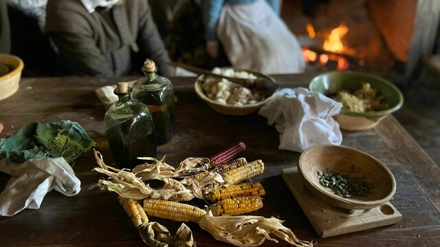 An array of food items and serve wear on a wooden table. Two pilgrims sit behind the table.