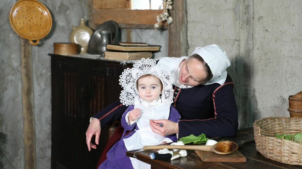 A mother pilgrim holds her infant daughter while seated at a kitchen table