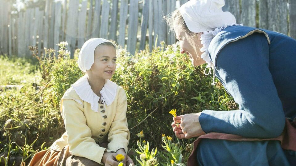 A pilgrim mother and daughter crouch in a garden while talking and picking flowers